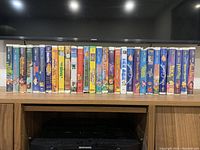 Front view showing 25 Disney VHS tapes lined up in a row with their spines visible on a wooden shelf beneath a TV.