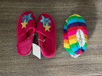 Two pairs of children's slippers; one pair red with star designs and Velcro strap, the other rainbow colored with plush lining.