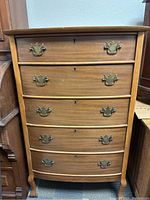 Front view of wooden chest of drawers showing the bowed front and five drawers with brass handles.