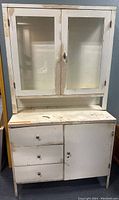 Front view of distressed white metal medical cabinet with two glass panel doors on top and three drawers plus one door on bottom.