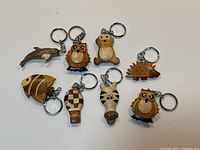 Photo showing seven wooden keychains including a dolphin, owl, bear, hedgehog, fish, hot air balloon with checkerboard pattern, and cow with metal key rings.
