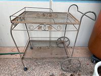 Metal outdoor 2-tiered cart showing both tiers, decorative sides, and wheels with one wheel loose.