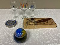 Overview of the lot showing 3 wine glasses, carving fork in box, Aspic Jelly Cutters tin, and glass paperweight on white patterned tablecloth