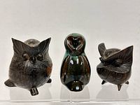 Front view of 3 owl figurines. Two are carved wood with carved eyes, ears and feathers, dark brown in color. One is a shiny glazed ceramic owl with smooth surfaces and green to black gradient color.