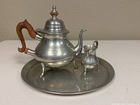 Full view of the pewter teapot, creamer, and tray set on a white surface with a beige wall background.