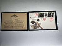 Open black presentation folder showing two coins mounted on holders and five Sherlock Holmes First Day Issue stamps canceled with red postmark. Printed text on left about Sherlock Holmes and his creator, printed image of magnifying glass and note on right.