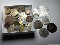 Photo of small white box containing variety of coins along with loose coins and coins in small plastic sleeves on white surface.