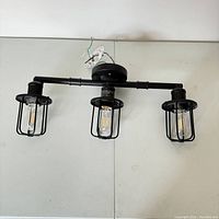 Front view of black adjustable vanity light fixture with three cage-enclosed bulbs spread across a horizontal bar, showing mounting base and wiring.