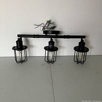 Front view of the black metal vanity light fixture with three caged bulbs, showing the horizontal bar and wiring.