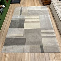 Top down image of the full rectangular area rug on wooden floor showing geometric blocks and stripes in neutral grey and taupe tones.
