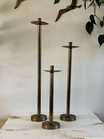Front view of set of three brass tone candle holders on a table against a wall, showing varying heights and slender stems.