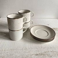 Set of four stacked off-white ceramic mugs with brown rims next to four matching saucer plates stacked on a white surface.