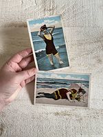 Two vintage swimming postcards: one propped vertically showing a woman standing in the ocean; the other horizontal showing a woman lying on the beach