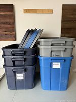 Stack of six Rubbermaid plastic storage bins with lids in black, blue, and gray colors, showing front view of stacked bins with labels