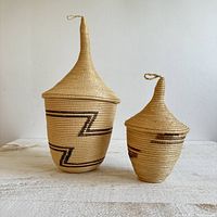 Side view of two neutral color African coil baskets with lids and pointed tops with loops, showing decorative zigzag patterns.