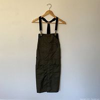 Full front view of olive green apron with adjustable black straps hanging on wooden hanger.
