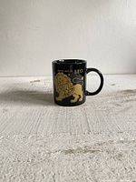 Black ceramic mug against a plain background showing the gold lion illustration and text 'LEO' and dates July 23 - August 22.