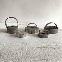 Front view of five vintage metal cookie cutters in assorted shapes and sizes with handles, arranged in a row on white textured surface.