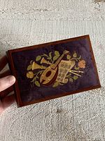 Top view showing wood framed rectangular music box with dark purple top featuring decorative music design including lute, trumpet, sheet music and floral vines.