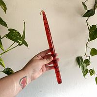 Photo showing hand holding vintage red Lucite tapered candle with foil flecks against a plain wall and green plant backdrop.