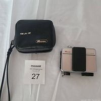 Photo of vintage Ricoh Flashmatic 500D camera and black leather case placed on white cloth background with auction tag showing "27".