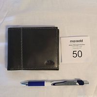 Photo of a black leather bifold wallet with white stitching and a small metal emblem, placed next to a pen and auction card for scale.