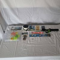 Wide shot showing the entire fishing gear lot laid out on white cloth. Includes rod and reel, spool of line, multiple packages of lures, baits and fishing tackle.