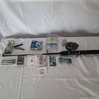 Fishing rod with attached black baitcasting reel and assorted fishing lures, tackle, and line laid out in a single line on white backdrop.