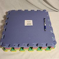 Stack of 8 foam puzzle piece flooring tiles in various colors, textured surfaces, interlocking edges, and a pen for scale next to them