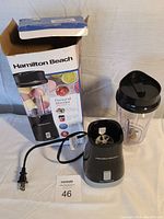 Photo showing the Hamilton Beach personal blender power base, clear blending cup with black lid, and original box which has wear.
