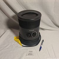 Front view of black Honeywell portable heater with two control knobs, showing attached yellow warning tag and power cord, placed on white surface.