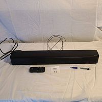 Full length front view of the LG sound bar, remote control, and pen for scale on a white cloth.