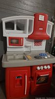 Front view of the Little Tikes play kitchen showing the red and gray plastic unit including microwave, stove, oven, sink, refrigerator, and vent hood. Missing faucet top piece.
