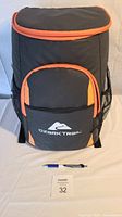 Black insulated cooler bag with orange trim, front zippered pocket and mesh side pockets, shown upright with pen for scale.