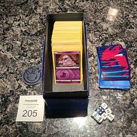 Shows a deck box containing a large stack of Pokemon cards with a purple Grimer card visible on top. Next to the box are a Pokemon coin and four multi-sided dice. To the right, there is a smaller stack of blue and red card sleeves.