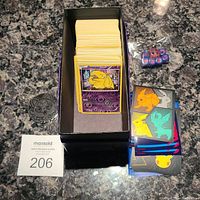 Top down view of the collection stacked in a black deck box, adjacent to assorted sleeves, coin, and dice on marble countertop.
