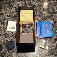 Full view of the lot containing a rectangular deck box filled with about 175 Pokémon cards, Pokémon-themed card sleeves stacked on the side, a Pokémon coin, and three numbered dice in a plastic bag.