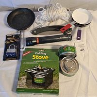 Photo of all camping supplies displayed on white surface including pans, rope, lantern, stove in box, can opener, cans, and padlock