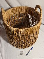 Photo showing round woven basket with thick braided weave, diamond pattern in middle, loop handles and pen for size reference.