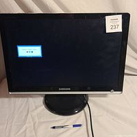 Samsung monitor powered on showing a message 'Check Signal Cable' on the screen, placed on white fabric surface with pen for scale.