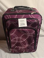 Small purple soft-sided suitcase with white circular line design, front zippered pocket, and black handle on top.