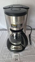 Front view of Waring coffee machine showing stainless steel body, glass carafe, digital control panel, and power cord.