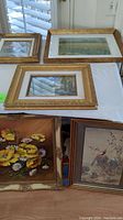 Five framed paintings on a table, two laid flat and three leaning against a wall. View shows variety of scenes and gold frames.