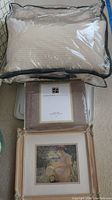 Beige pillows in clear plastic carry bag with black trim on top, below are packaged beige full size sheet set and framed art print.