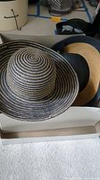 Photo of multiple hats including a black and beige spiral straw sun hat with wide brim and a tan and blue wide brim hat in a box.