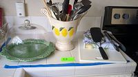 Wide shot showing ceramic utensil holder with lemon design, glass dome cake plate, green decorative plate, folded lemon-patterned cloths, and kitchen knives on white tiled counter.