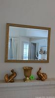 Gold-framed mirror mounted above a white brick mantel, with two gold bird figurines and gold vase placed on the mantel below.