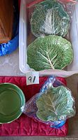 Four leaf-shaped ceramic plates stored in a plastic bin and on a red surface; two light green cabbage leaf plates and two darker green textured cabbage leaf plates