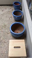 Three blue pottery pots of different sizes arranged in a row on a patio floor, showing their open tops and glazed blue finish.