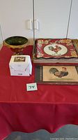 Ceramic rooster serving tray, two placemats with rooster images, green pedestal bowl, and two boxed candles set on a red cloth.